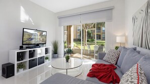 A modern living room with a flat-screen TV, a glass coffee table, a grey sofa, and a red throw blanket. - One in a Minion themed condo, 5 mins to Disney | WiFi + Keurig (Kissimmee)