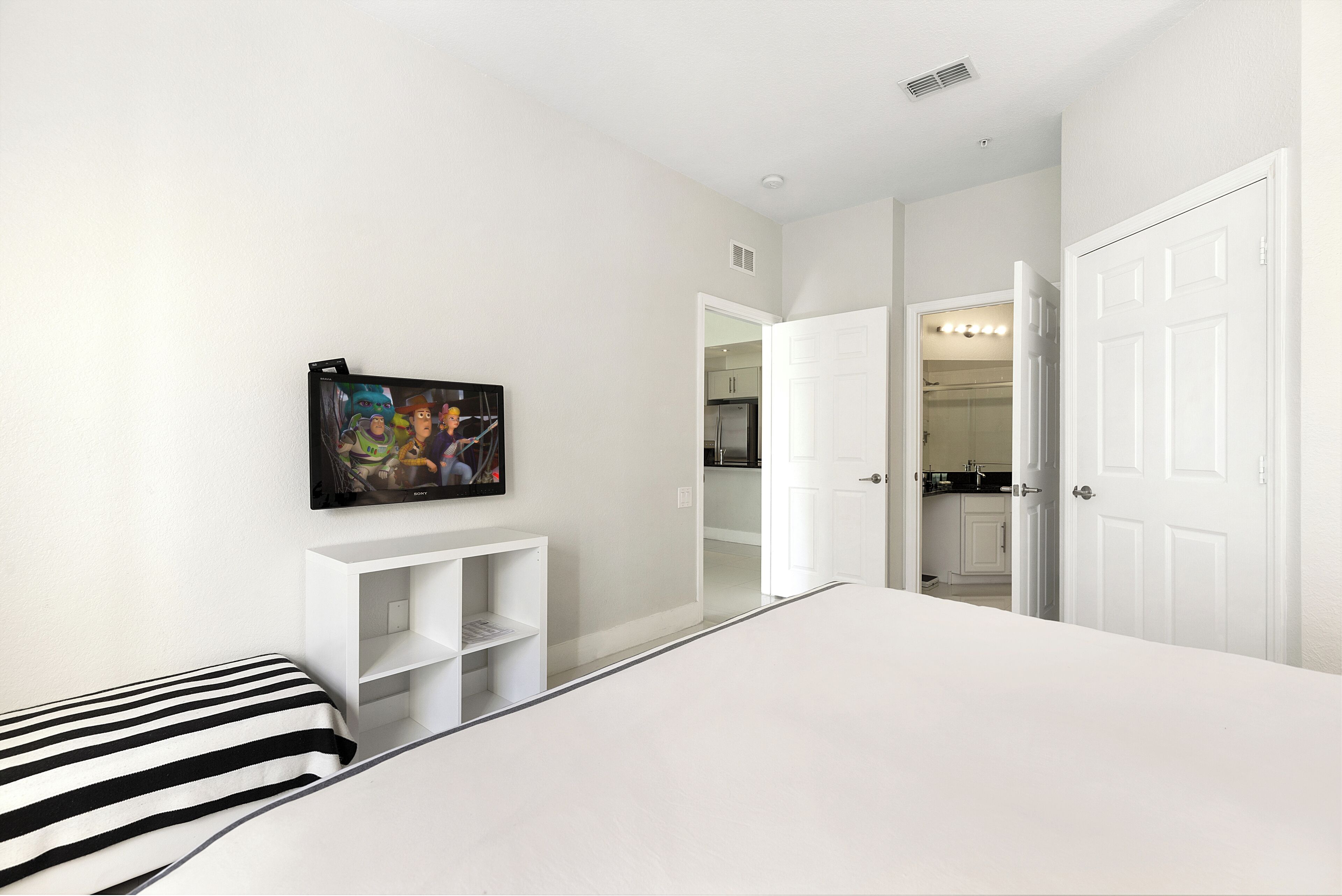 A bedroom with a bed, a TV mounted on the wall, a white wardrobe, and a door leading to a bathroom.