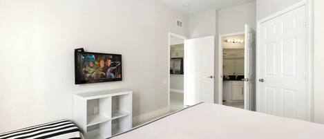 A bedroom with a bed, a TV mounted on the wall, a white wardrobe, and a door leading to a bathroom.