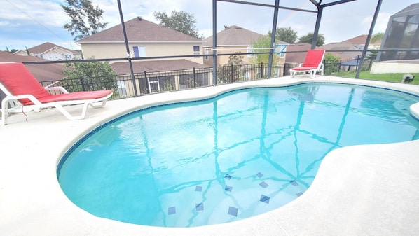 Una piscina con bordo piastrellato, circondata da un solarium bianco. Si vedono due sdraio rosse e un recinto metallico sullo sfondo.
