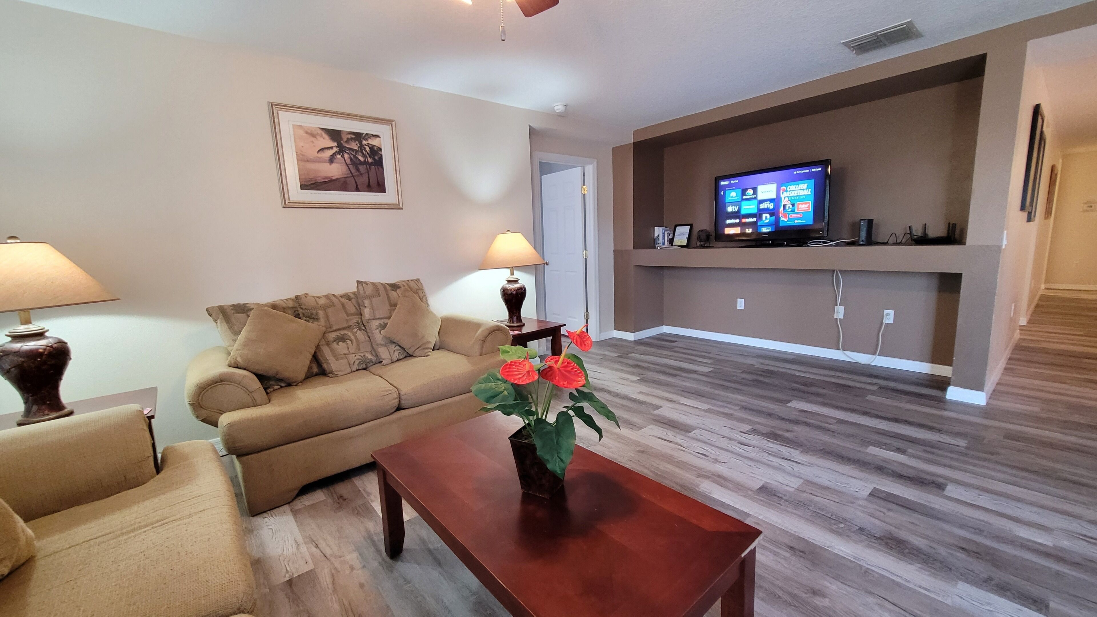 A living room with a sofa, two lamps, a coffee table with flowers, a TV on a stand, and a door leading to another room.