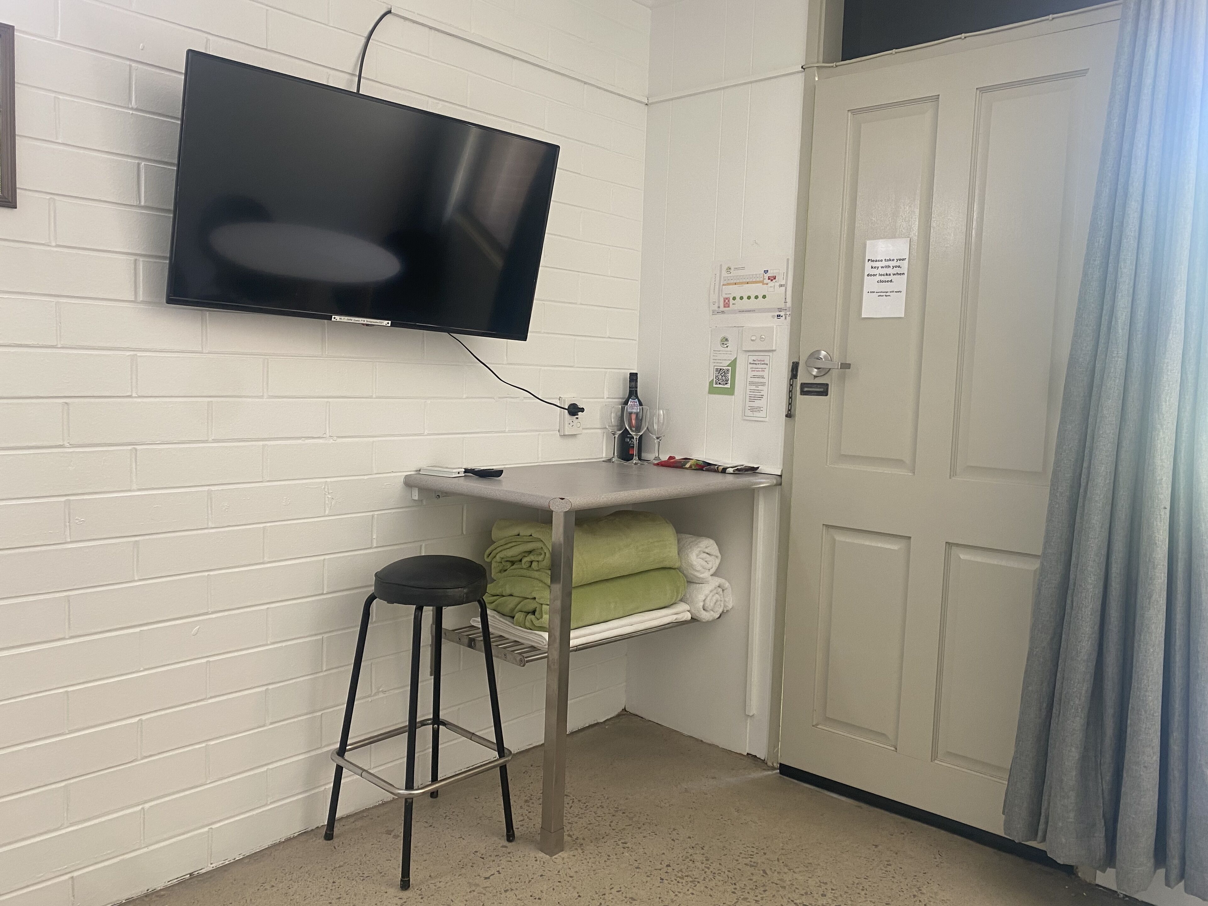 A room with a flat-screen TV mounted on the wall, a small bar counter with a stool, a door with a sign, and a towel rack with towels.