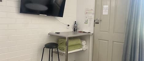 Una habitación con un televisor de pantalla plana montado en la pared, una pequeña barra con un taburete, una puerta con un cartel y un perchero con toallas.