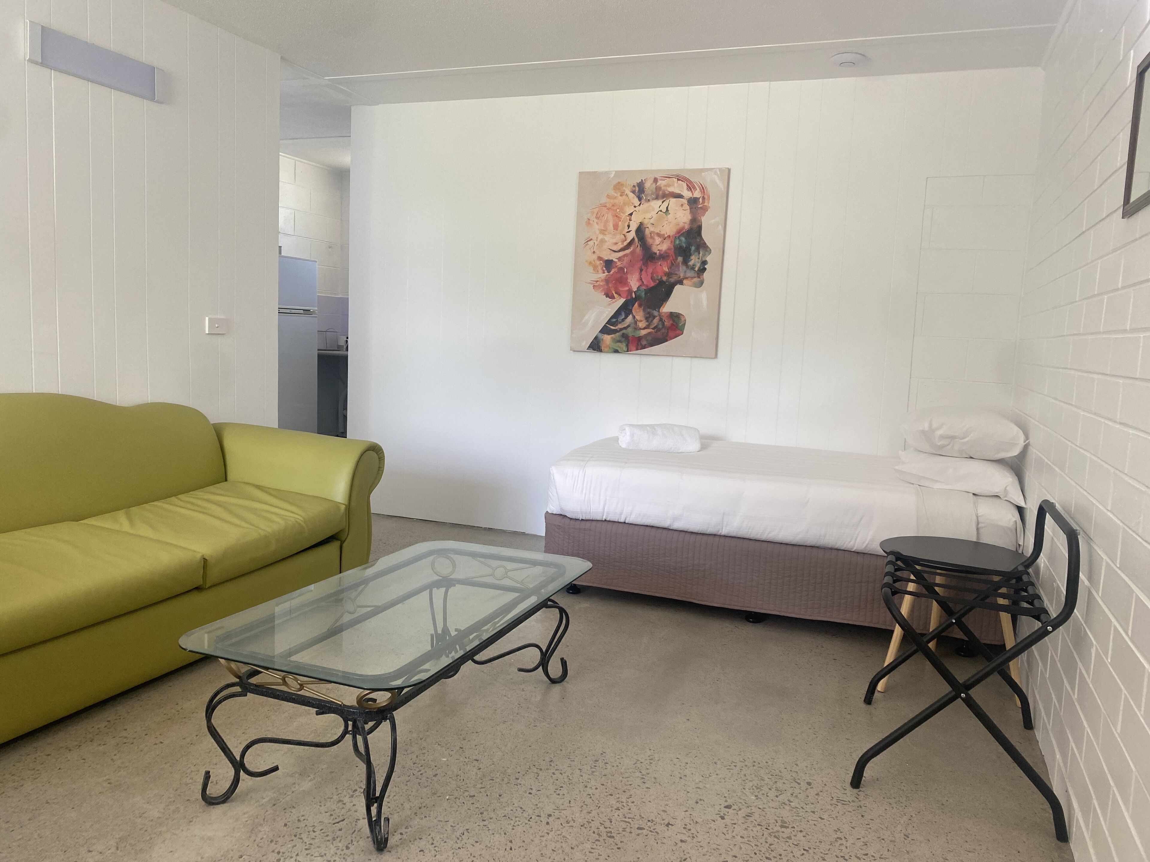A room with a green sofa, a glass-top table, a bed with white bedding, and a painting on the wall.