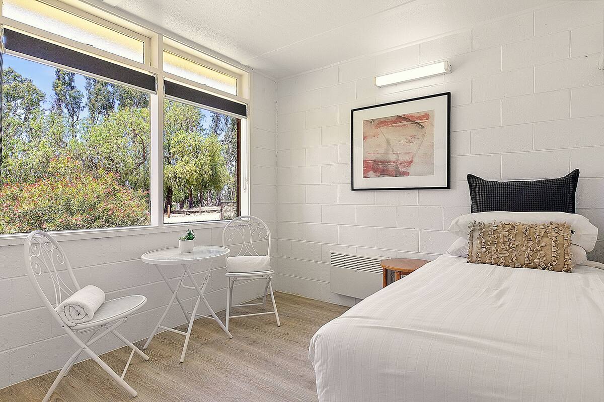 A bedroom with a bed, a chair, a small table, and a window with a view of trees.