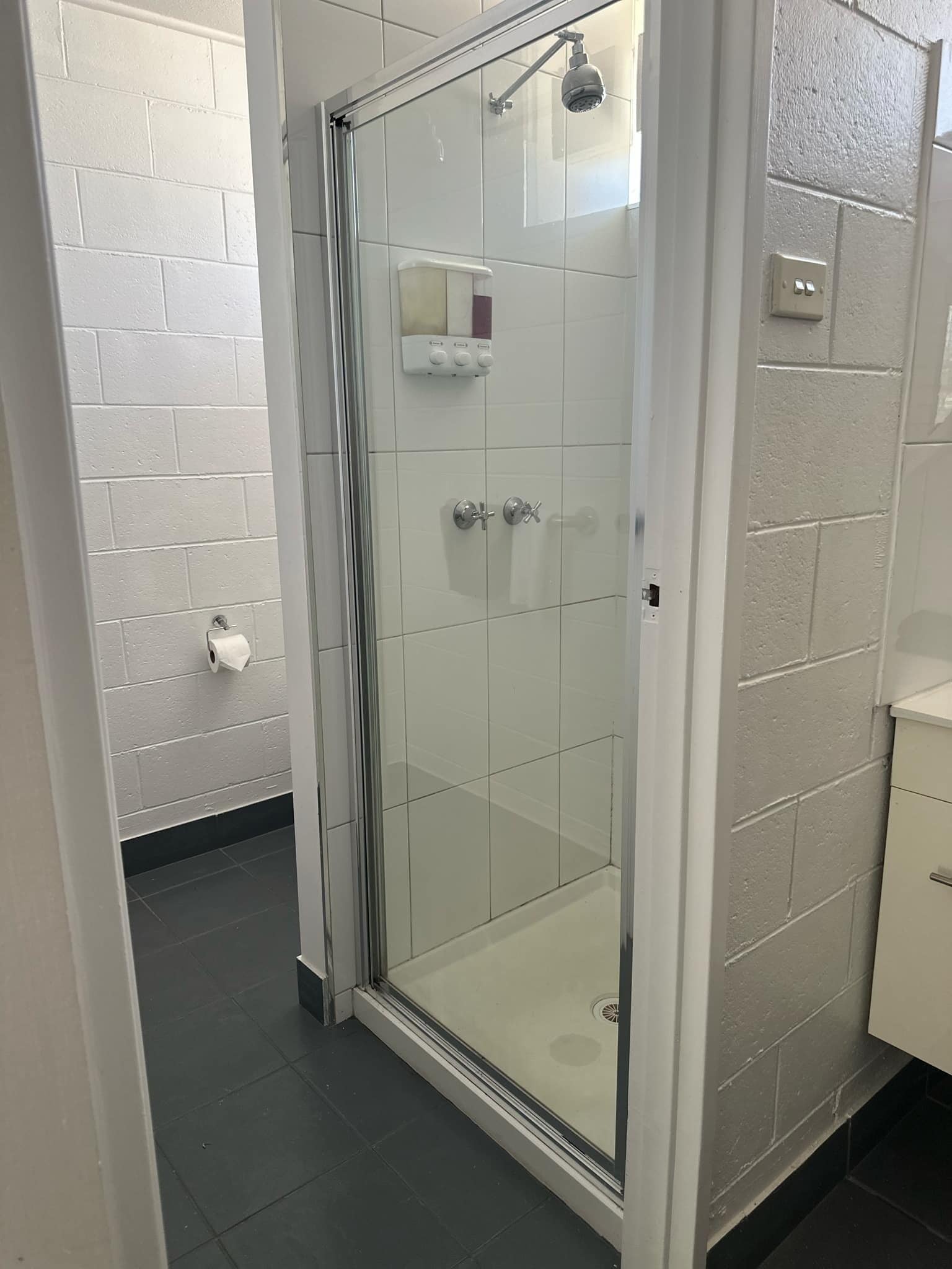 Une cabine de douche fermée par des parois en verre, avec un mur recouvert de carreaux blancs, une porte blanche et une petite étagère contenant des articles de toilette.