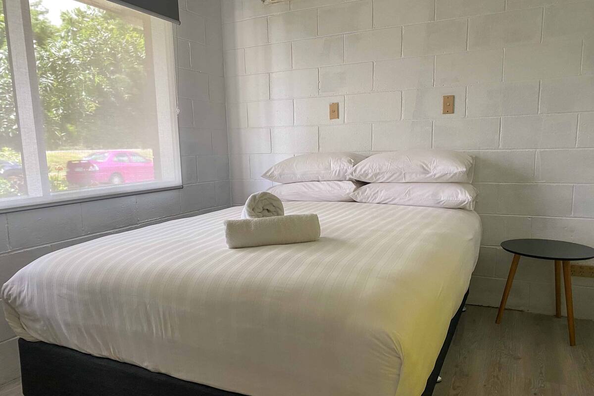 A neatly made bed with white linens and a rolled towel, a small round table, and a window with a view of trees and a car outside.