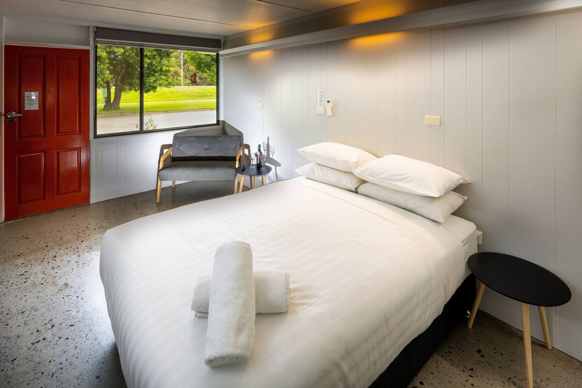 A hotel room with a bed, a small table, a chair, and a window with a view of greenery.