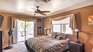 A bedroom with a large bed, a ceiling fan, a standing fan, a nightstand, and a window with curtains.