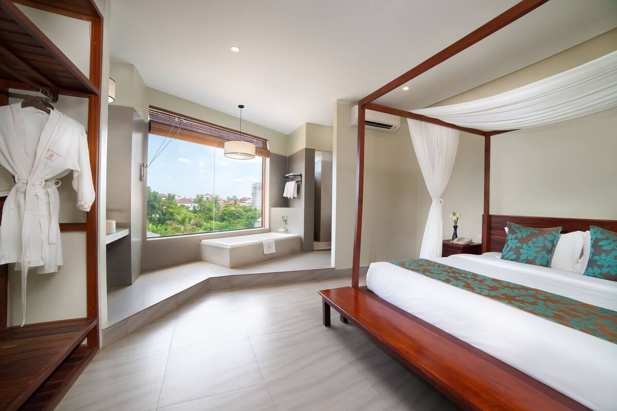 A modern bedroom with a four-poster bed, a bench, a vanity, and a window with a view of greenery.