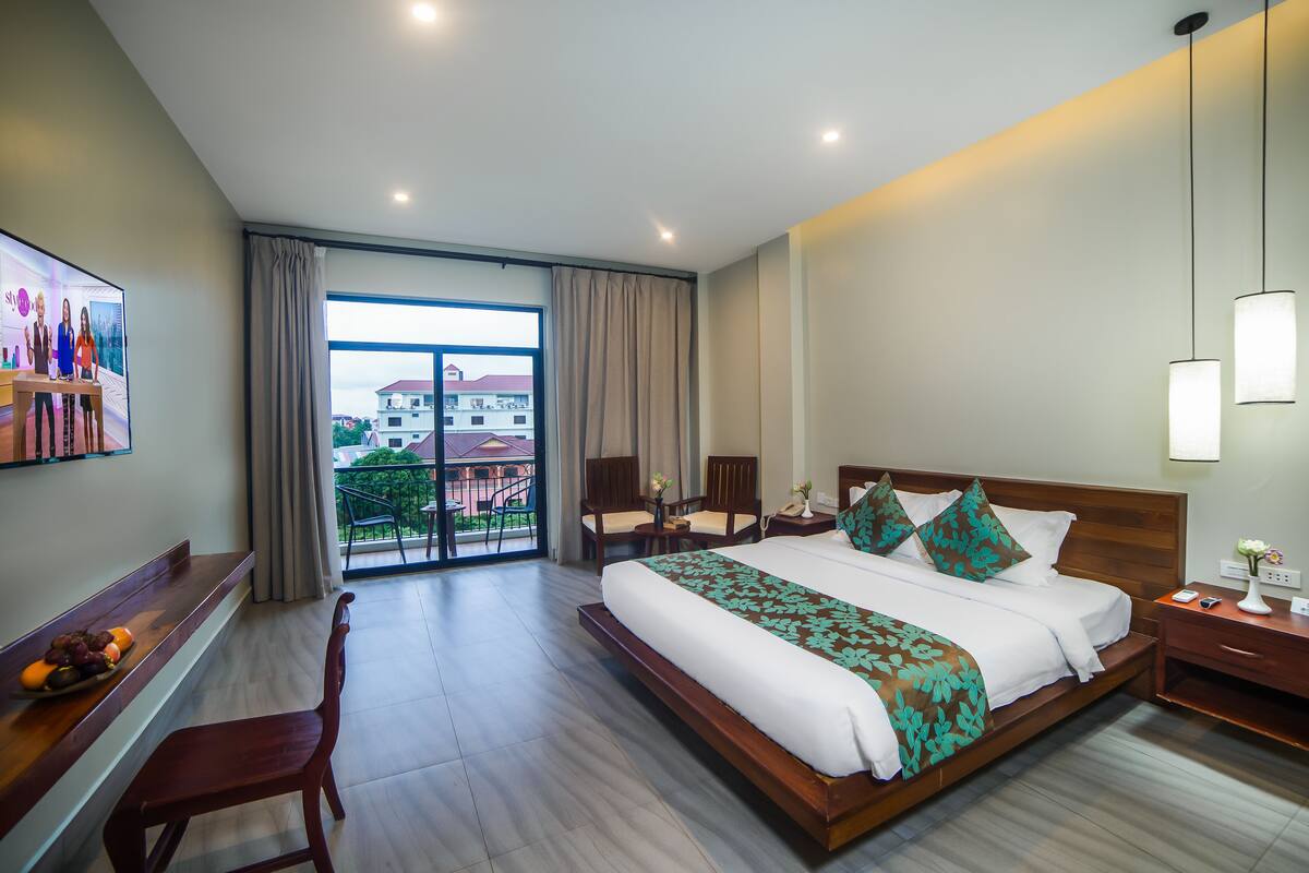 A hotel room with a large bed, a desk with a chair, a TV, and a balcony with a view of buildings.