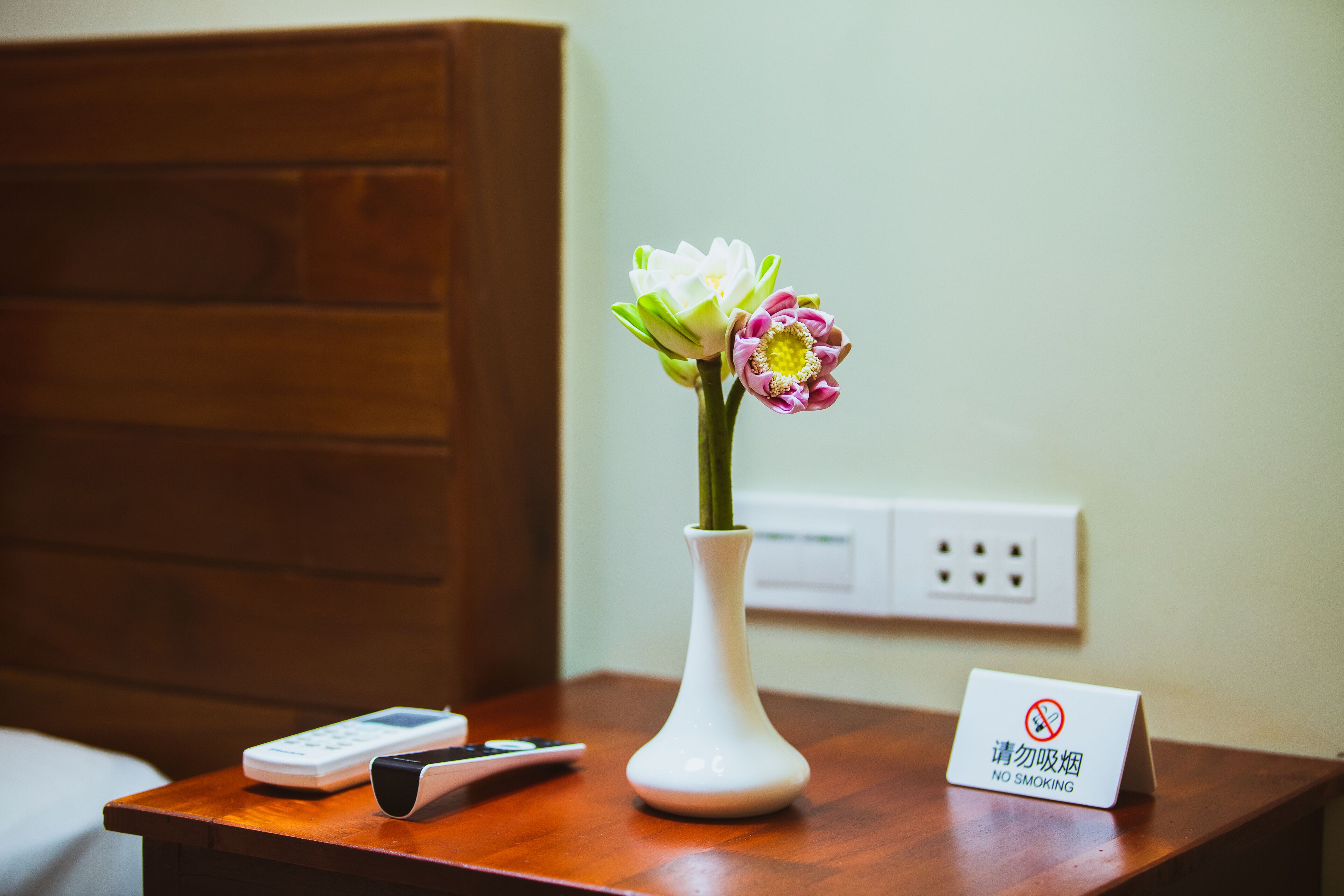 Ett träbord som står vid sängen, med en vit vas som innehåller blommor, en fjärrkontroll, en telefon och en skylt med texten ”Rökning förbjuden”.