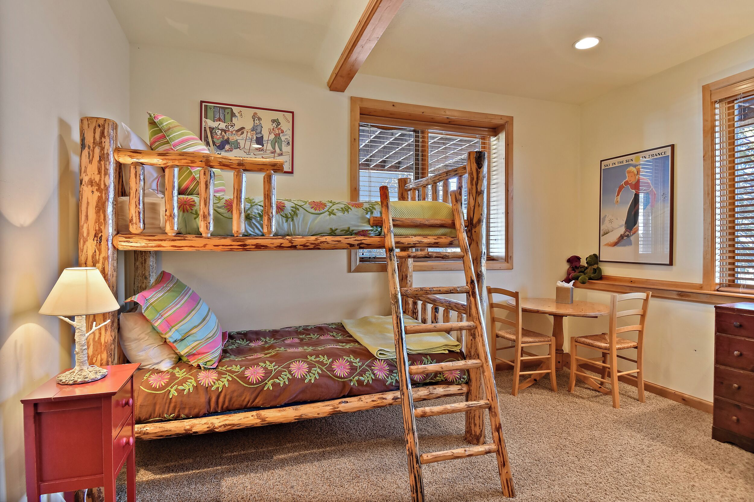 A bunk bed with a wooden ladder, a desk with chairs, a lamp, and a window with blinds.
