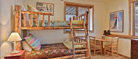 A bunk bed with a wooden ladder, a desk with chairs, a lamp, and a window with blinds.