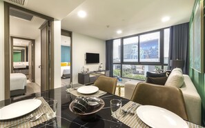 Une chambre d’hôtel moderne avec un coin repas, un lit avec des oreillers jaunes, une télévision et une vue sur la ville.