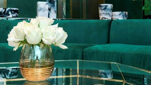 A green sofa, a glass coffee table with a vase of white flowers, and marble vases on shelves in the background. - Maven Stylish Hotel Bangkok (Bangkok)