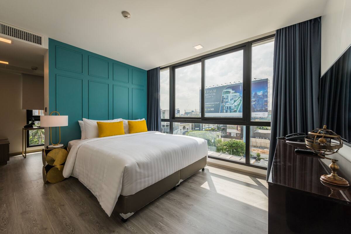 Ein modernes Hotelzimmer mit einem großen Bett, Akzentwänden in Türkis, Blick auf die Stadt und einem Schreibtisch mit Lampe.