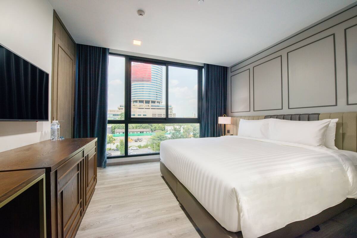 Ein Hotelzimmer mit einem großen Bett, einem hölzernen Schminktisch, einem Flachbildfernseher und einem Fenster mit Blick auf die Stadt.