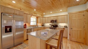 A kitchen with wooden cabinets, a stainless steel refrigerator, granite countertops, and a central island with bar stools. - Maggie Valley Cabin w/ Private Hot Tub & Game Room (Maggie Valley)