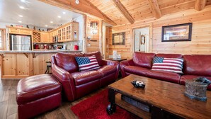 A cozy cabin interior with a wooden ceiling, leather sofas with American flag pillows, a wooden coffee table, and a kitchen area with stainless steel appliances. - Catalina Creekside Cabin - Log Cabin, Perfect for Couples, Fenced Yard (Big Bear Lake)