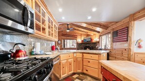 A modern kitchen with wooden cabinets, granite countertops, a gas stove, and a stone fireplace. - Catalina Creekside Cabin - Log Cabin, Perfect for Couples, Fenced Yard (Big Bear Lake)