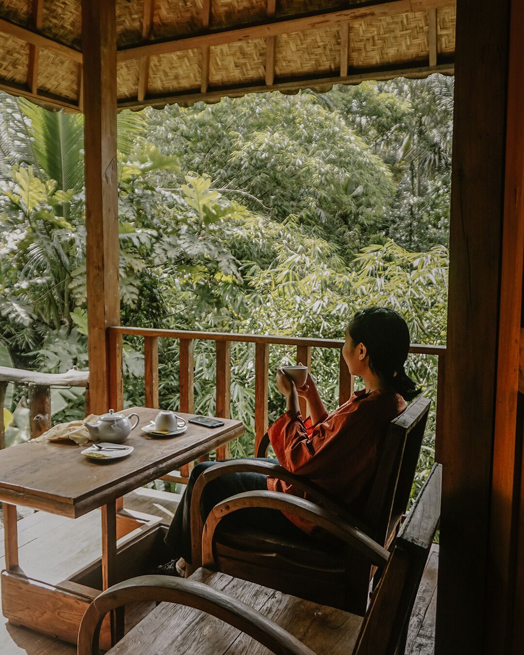 A person seated at a wooden table with a teapot and cups, overlooking a lush garden.