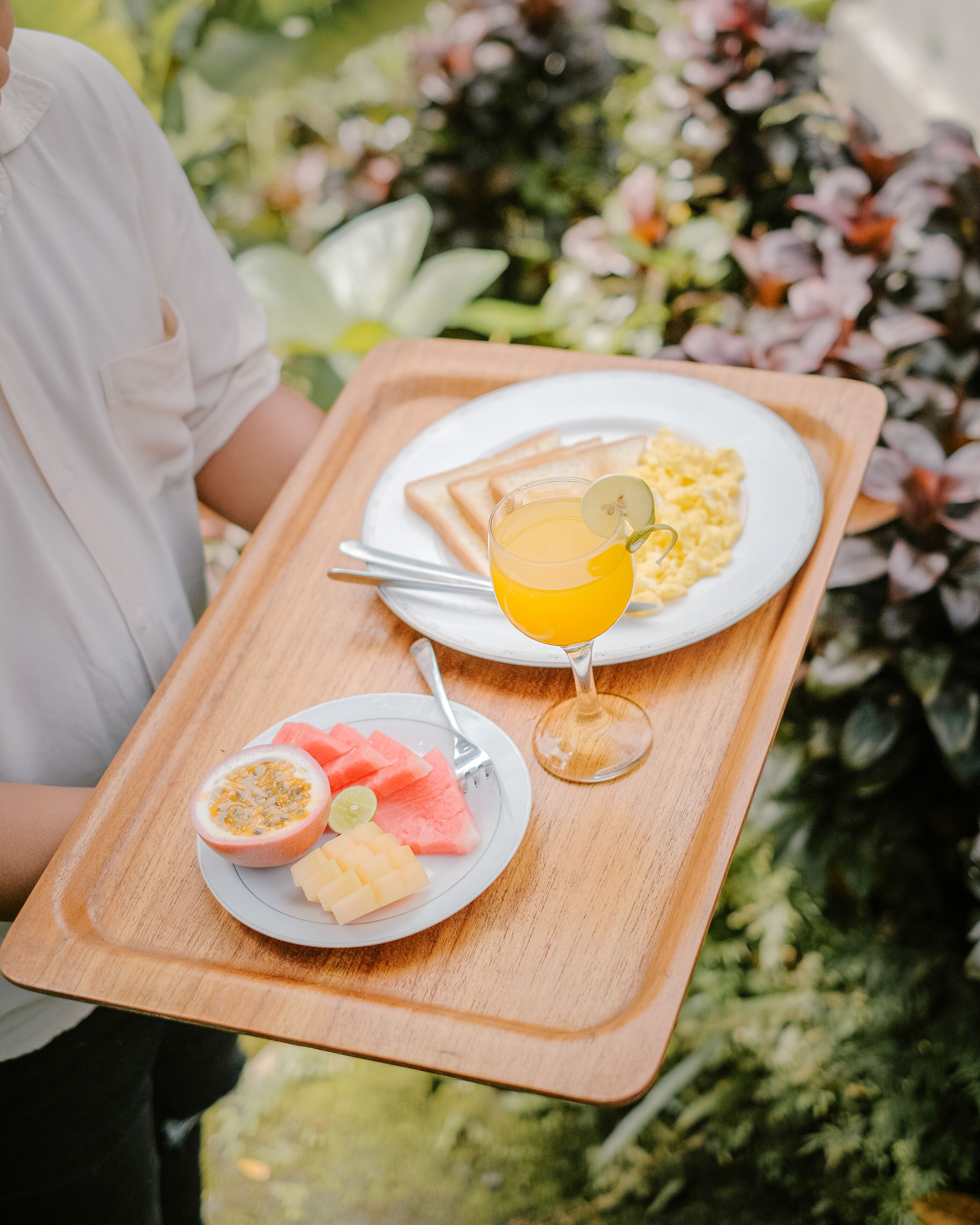A wooden tray with breakfast items including toast, scrambled eggs, a glass of orange juice, watermelon, and passion fruit.