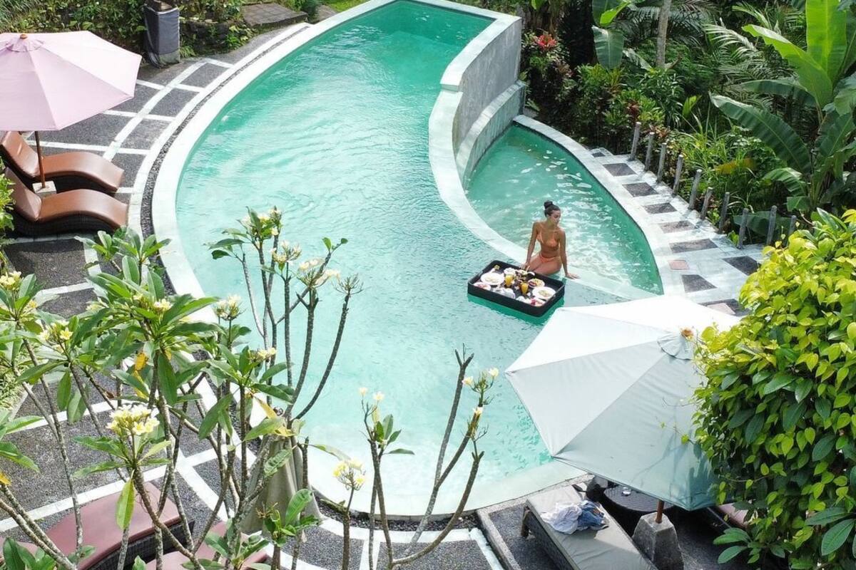 A pool with a curved edge, surrounded by lounge chairs and umbrellas, with a person sitting on a floating platform.