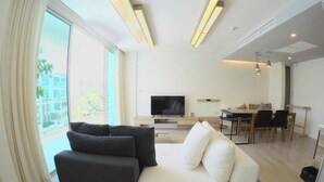 A modern living room with a sofa, a flat-screen TV, a dining area, and a large window with curtains. - Luxury Wanvayla Condo (Hua Hin)