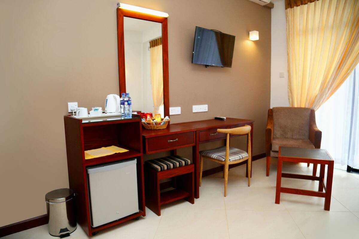 A hotel room with a wooden desk, a chair, a small refrigerator, a mirror, a television, and a window with curtains.