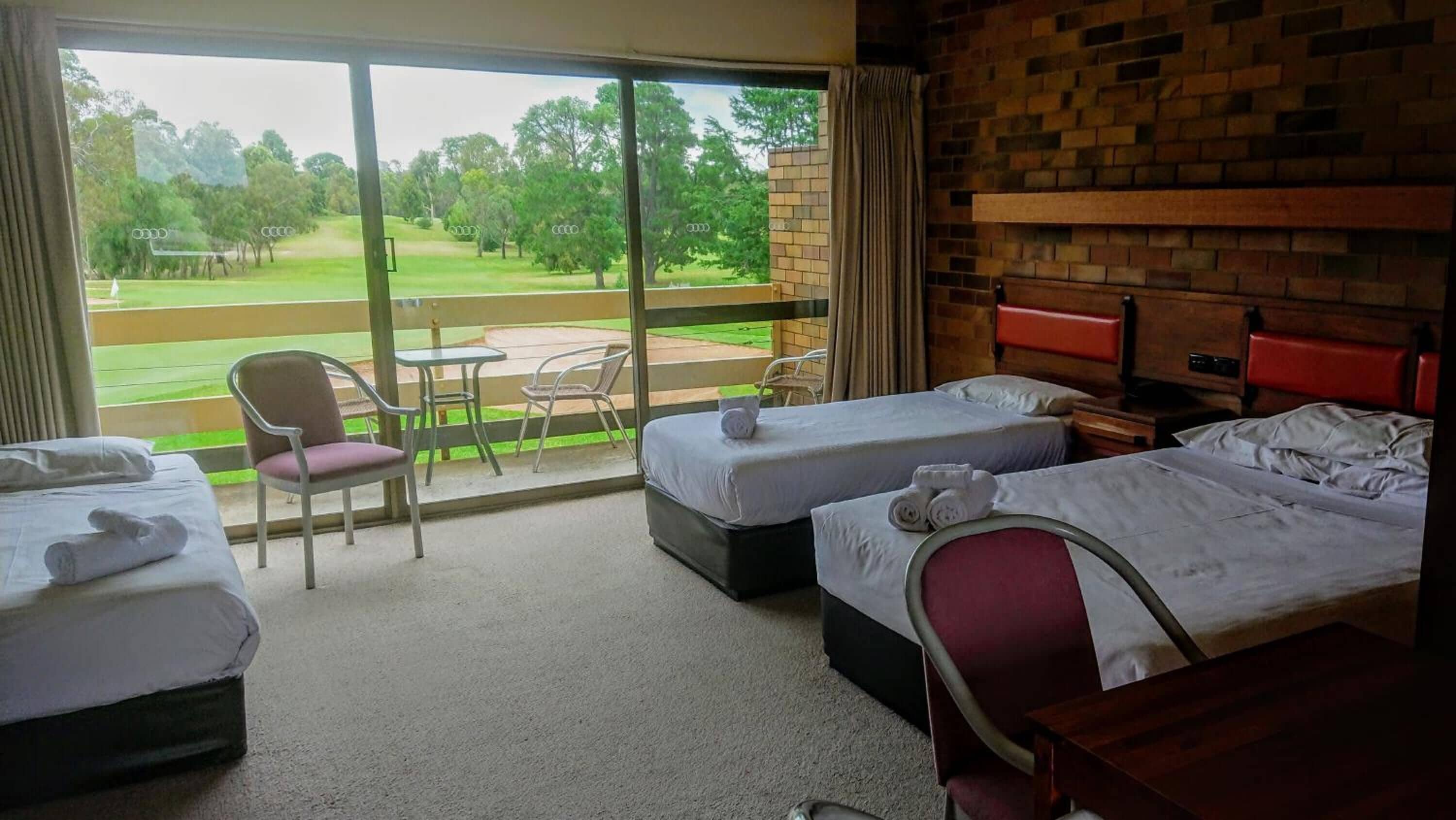 A hotel room with two beds, a chair, a small table, and a view of a golf course through large windows.