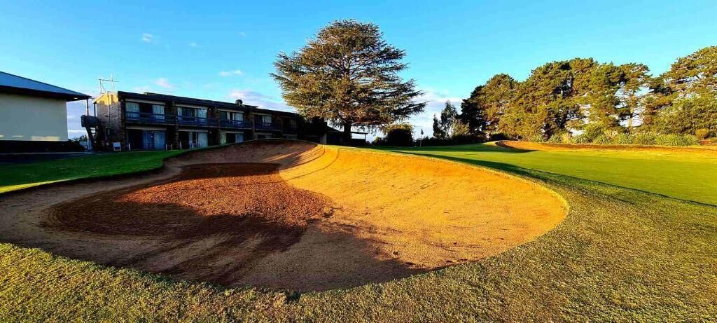 Ein Golfplatz mit einem Bunkersand, einem mehrstöckigen Gebäude und einem großen Baum.