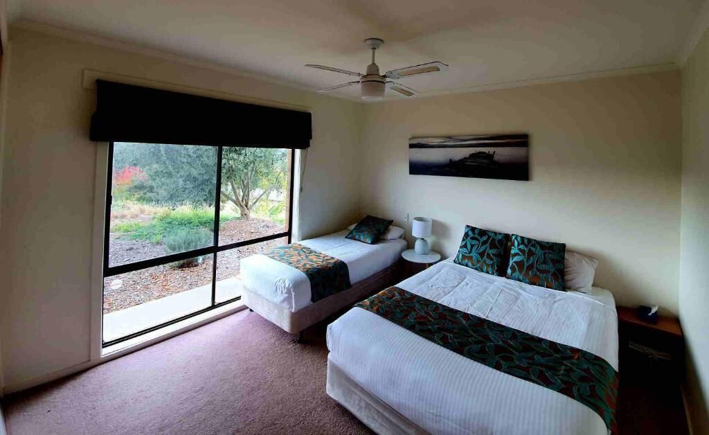 A bedroom with two beds, a ceiling fan, a window with a view of greenery, and a painting on the wall.