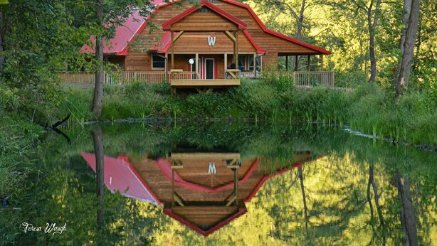 A wooden cabin with a red roof, surrounded by lush greenery and a calm lake.