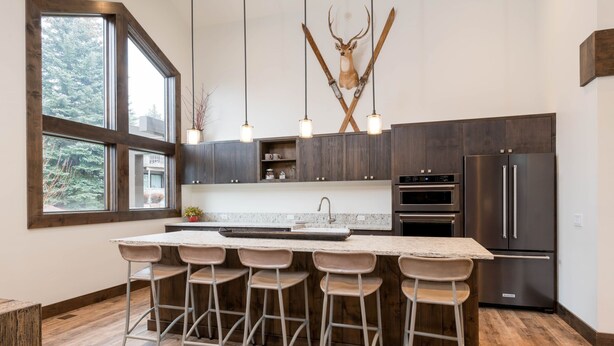 A modern kitchen with a large island, bar stools, dark wood cabinets, stainless steel appliances, and a deer head mounted on the wall. - Book A Getaway Today! Updated Corner Condo; Close to Dollar Mountain (Sun Valley)