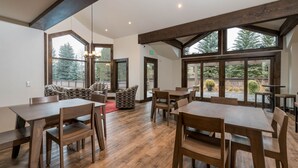 A spacious dining area with wooden tables and chairs, large windows offering views of trees, and a cozy seating area with patterned cushions. - Book A Getaway Today! Updated Corner Condo; Close to Dollar Mountain (Sun Valley)