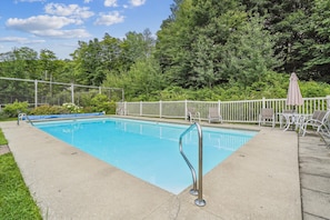 A rectangular swimming pool with a metal ladder, surrounded by a white fence, and equipped with lounge chairs and an umbrella.