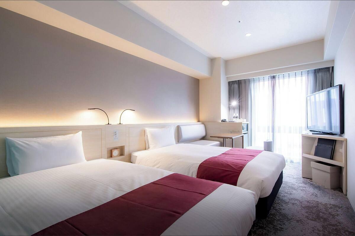 Ein Hotelzimmer mit zwei Betten, einem Schreibtisch, einem Fernseher und einem Fenster mit Vorhängen.