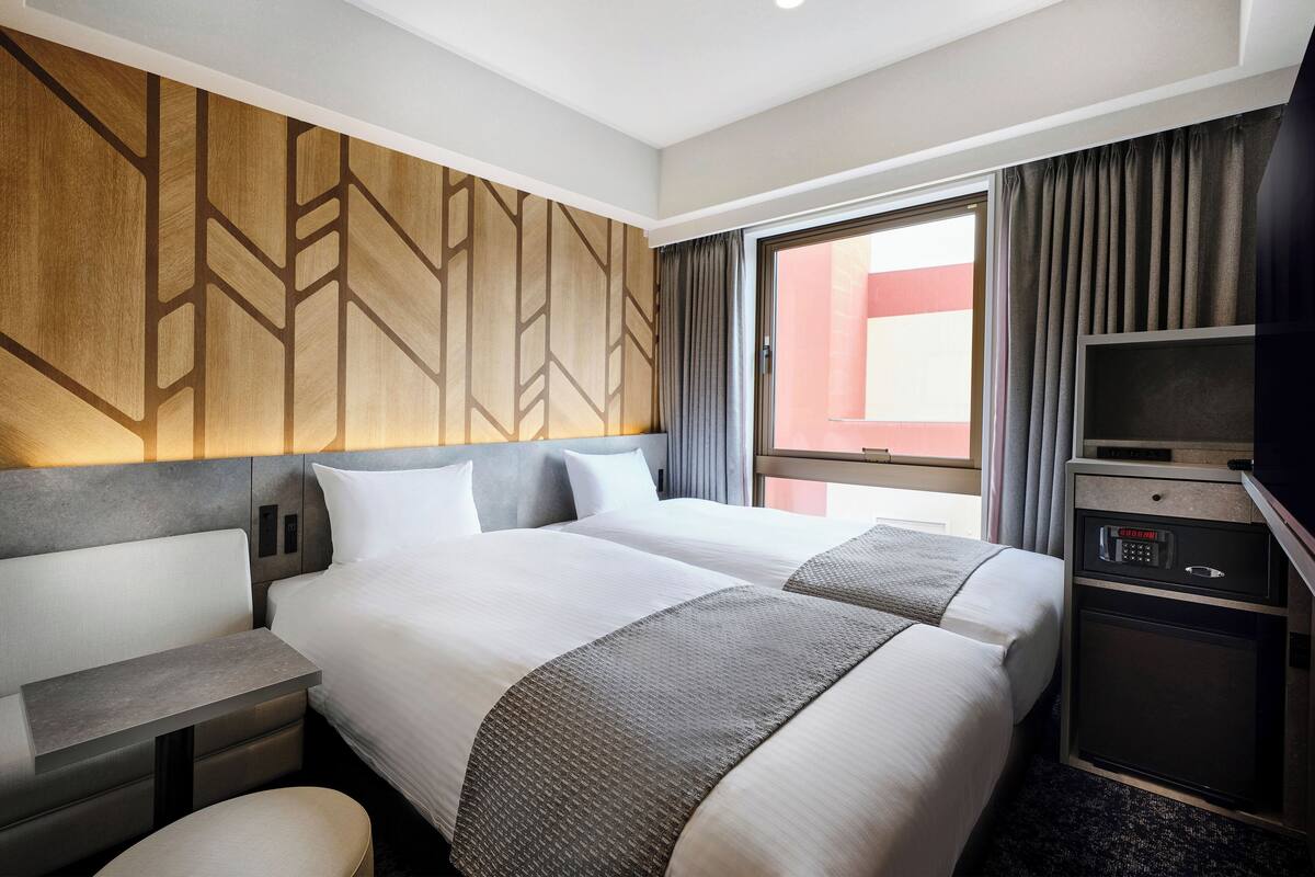 Ein Hotelzimmer mit zwei Betten, einem kleinen Schreibtisch, einem Fernseher und einem großen Fenster mit Vorhängen.