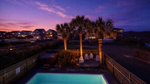 A residential area with a swimming pool, palm trees, and houses with illuminated windows. - Island Time - NH MP 11, 50 Ft to Beach, Elevator, Pool, Free Pool Heat, Hot Tub, Pets OK, YR Flex (Nags Head)