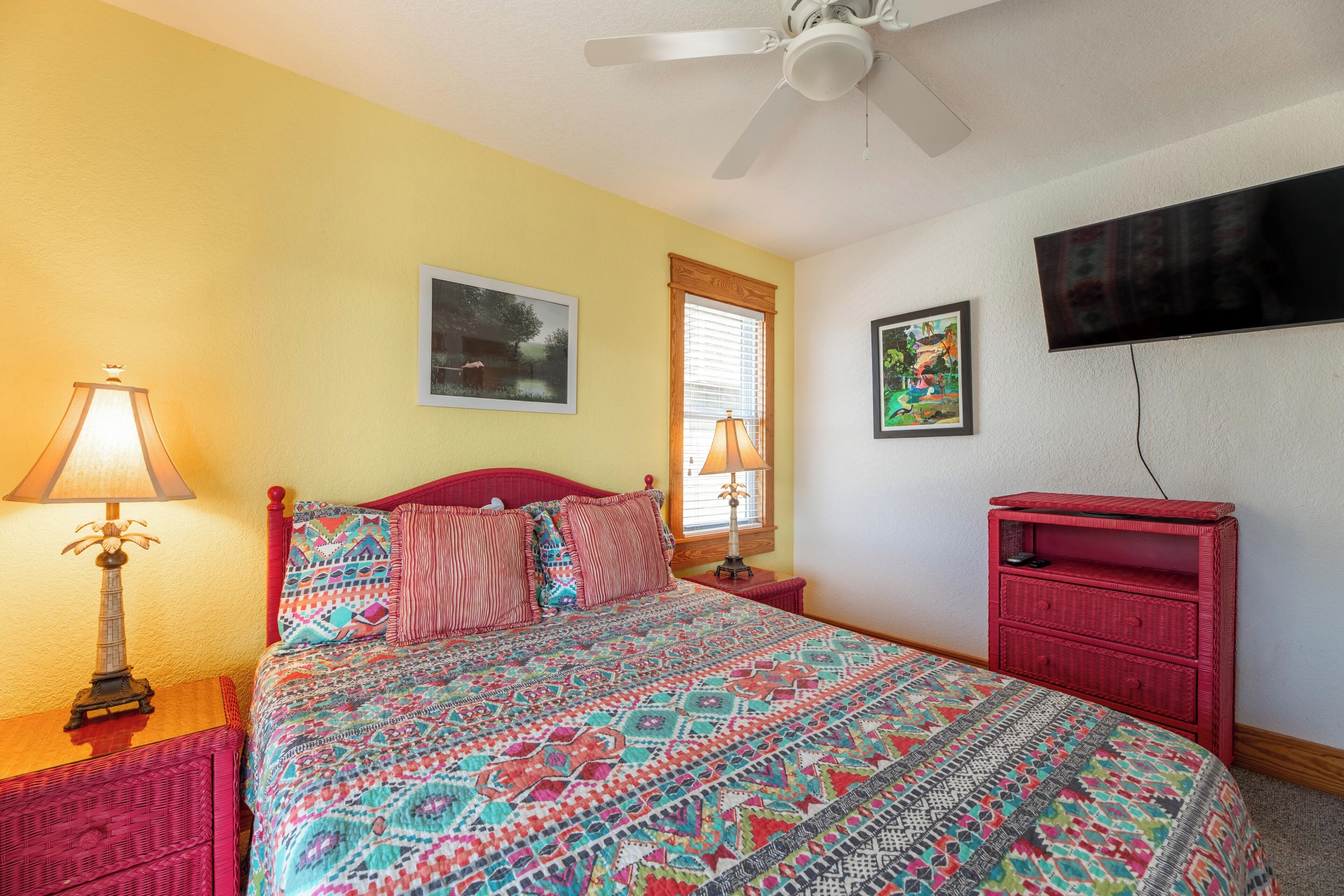 A bedroom with a bed, two bedside lamps, a ceiling fan, a TV mounted on the wall, and a red nightstand.