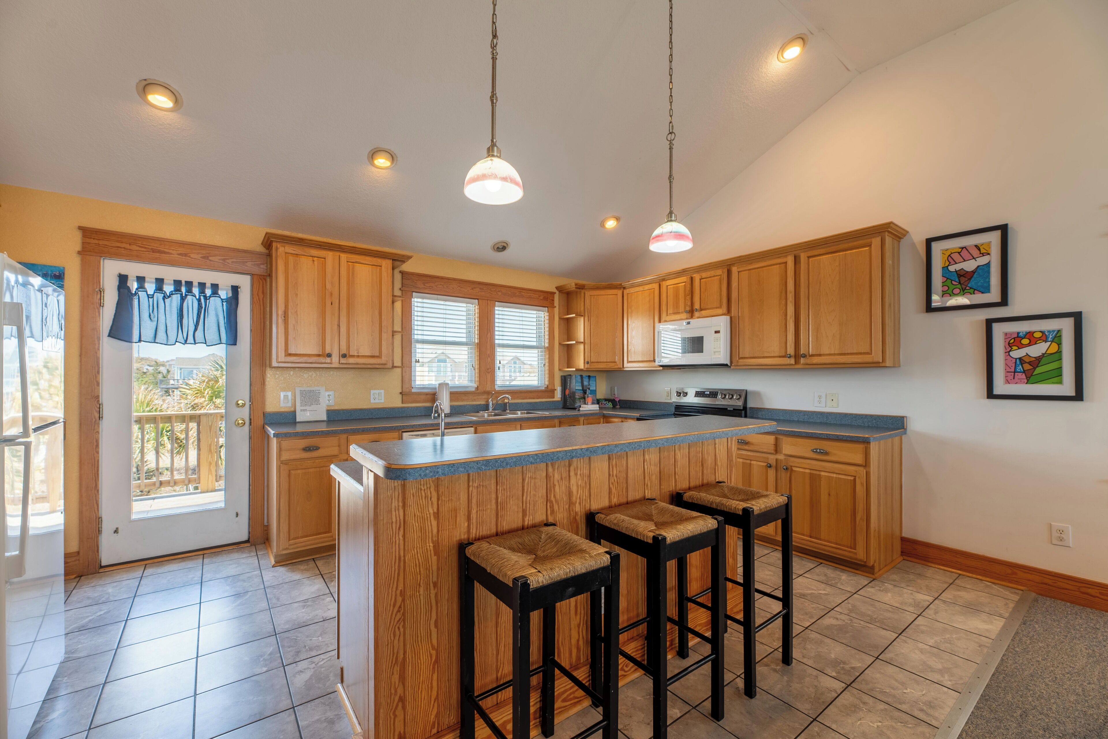 A kitchen with wooden cabinets, a large island with stools, a microwave, and pendant lighting.