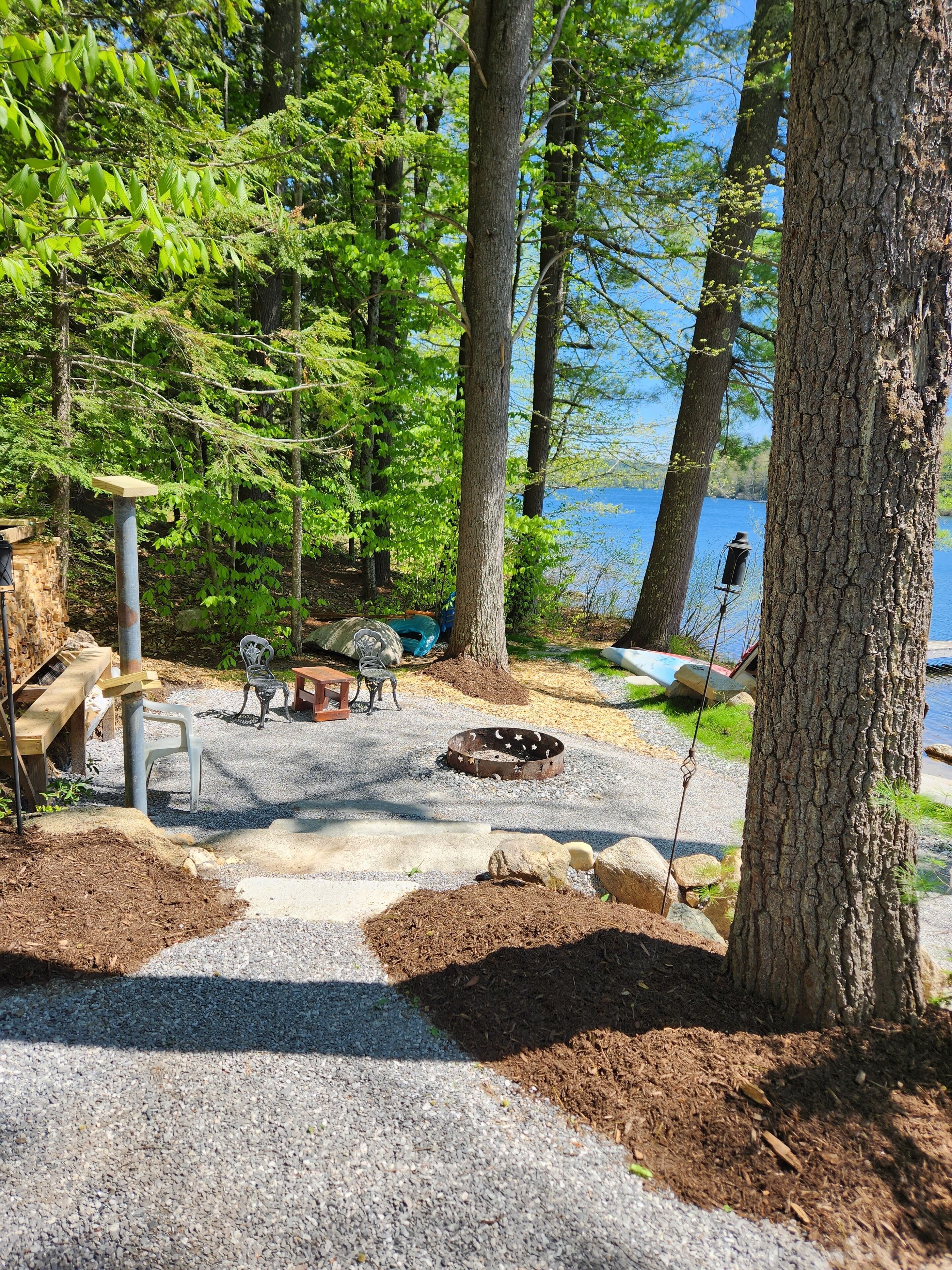Un camping paisible avec un foyer, une table de pique-nique et des bancs, entouré d’arbres et offrant une vue sur un lac.
