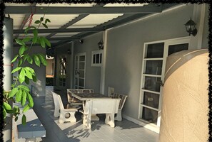 Uma área de varanda coberta com mesa, cadeiras, um vaso de planta e uma janela grande.