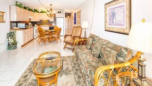 Amplia sala de estar con un sofá, sillas y una mesa de centro. Se observa un área de cocina con armarios y una mesa de comedor.