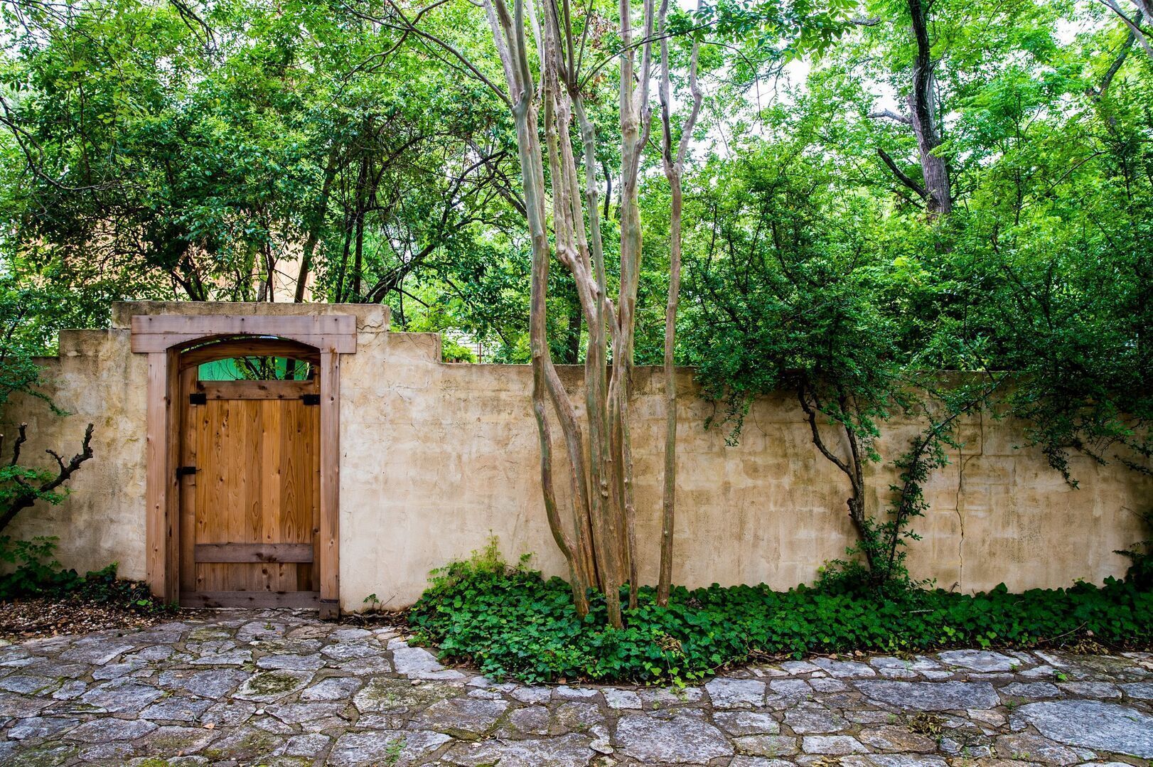 Una puerta de madera rústica da paso a un camino de piedra rodeado de exuberante vegetación y un muro de piedra.