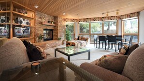 Un salon chaleureux avec une cheminée, des sièges confortables et de grandes fenêtres donnant sur l’extérieur.