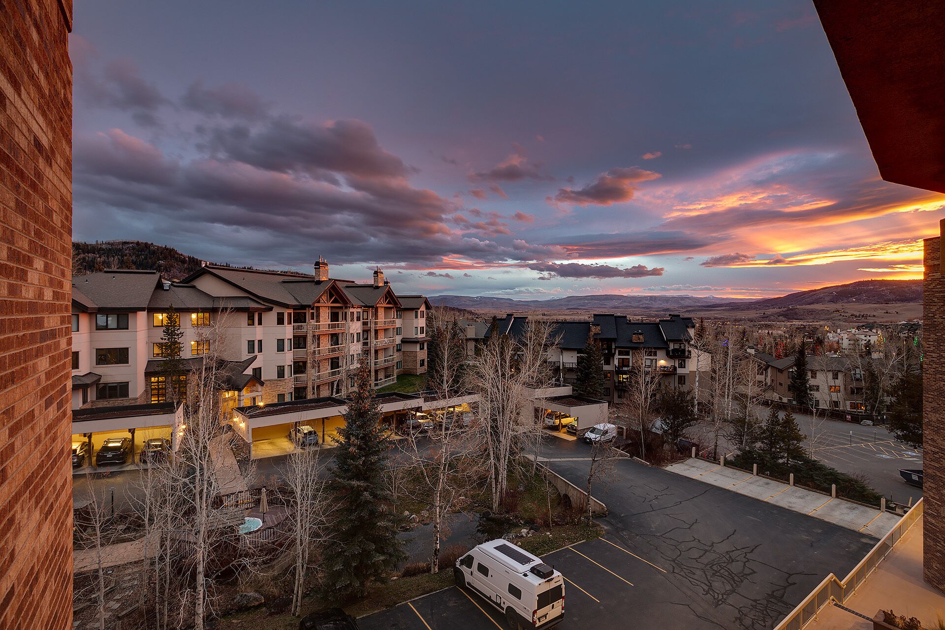 A multi-story hotel with a parking area, surrounded by trees and a view of the mountains.