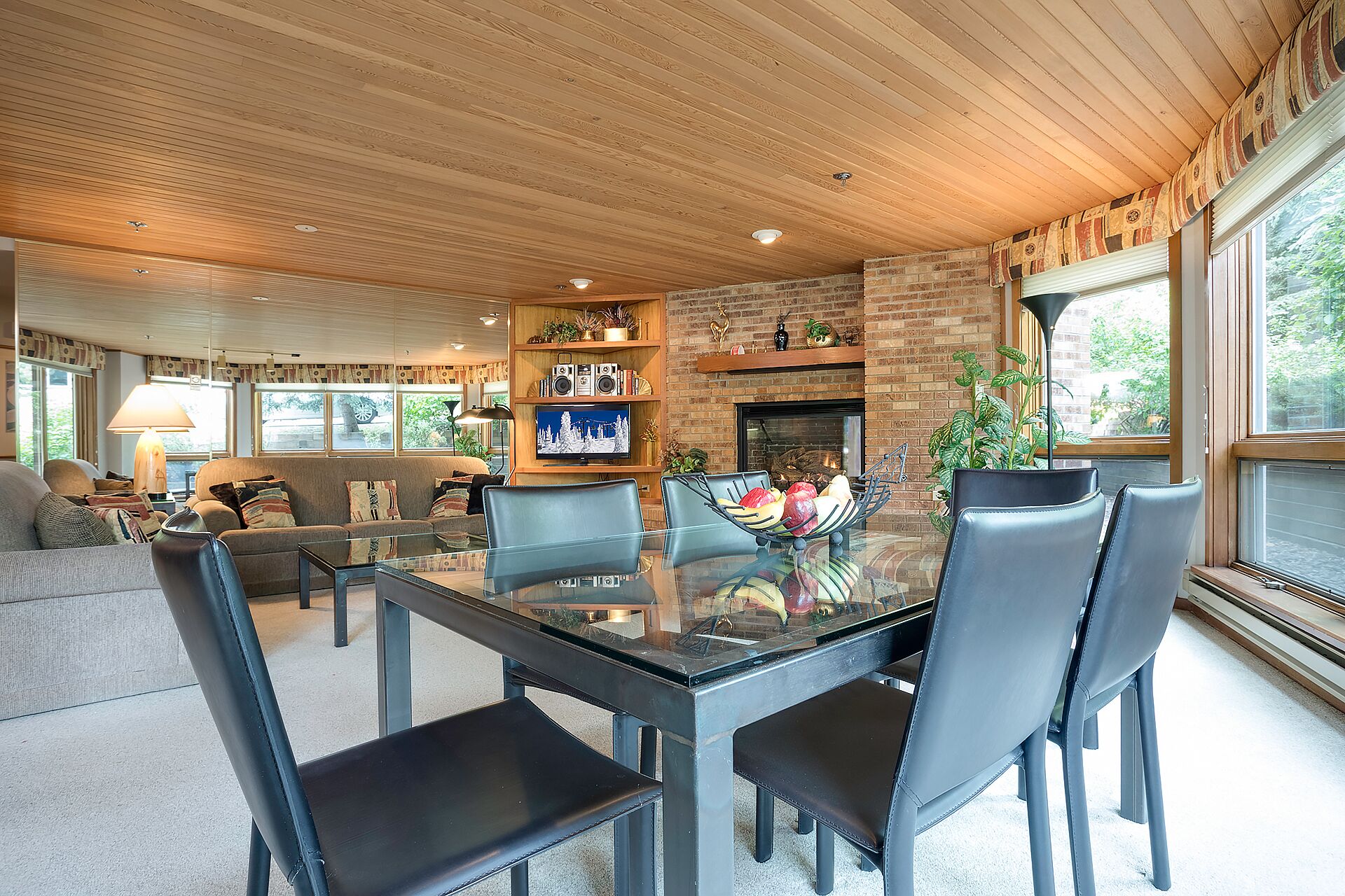 Un salon spacieux avec une table à manger au plateau en verre, des fauteuils en cuir, une cheminée en briques et une grande fenêtre donnant sur l’extérieur.