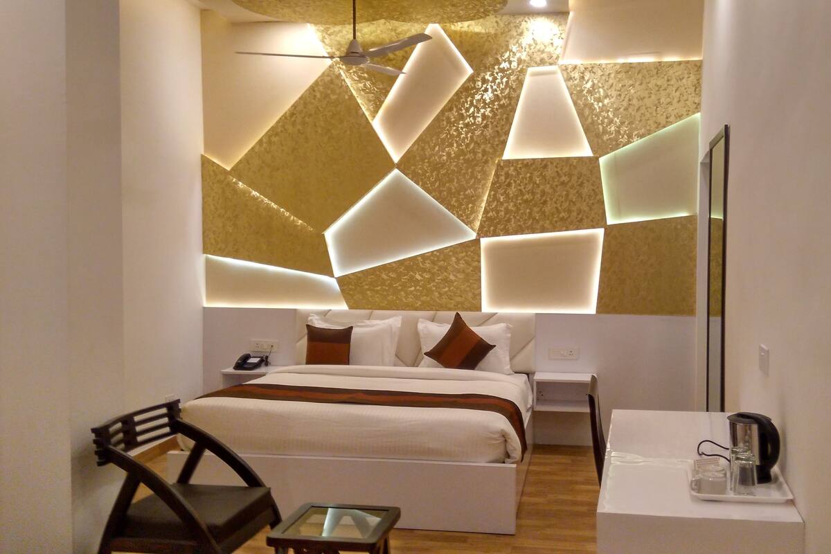 A modern hotel room with a bed, bedside table, chair, and a small table with a kettle and cups.